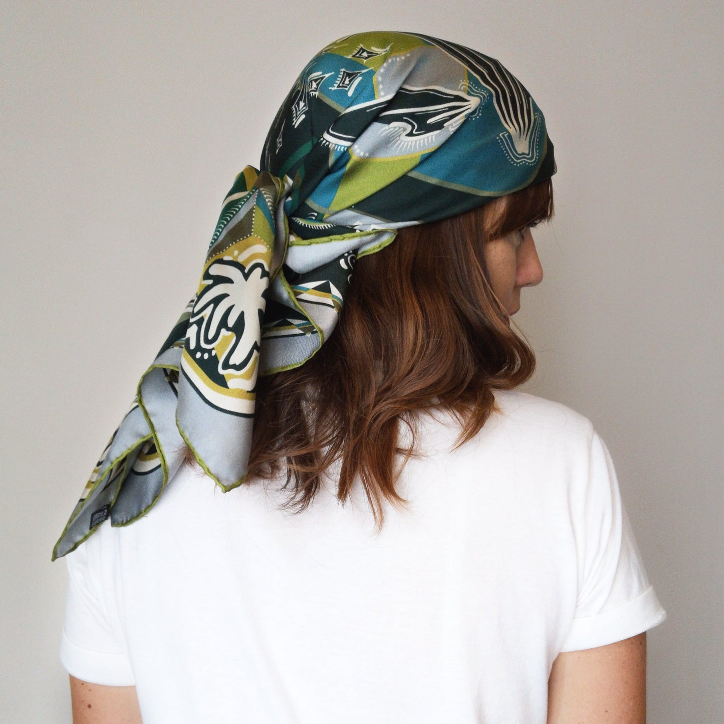 foulard di seta carré indossato in testa vista retro annodato stile bandana colori verdi ottanio ocra e avorio e disegno illustrato