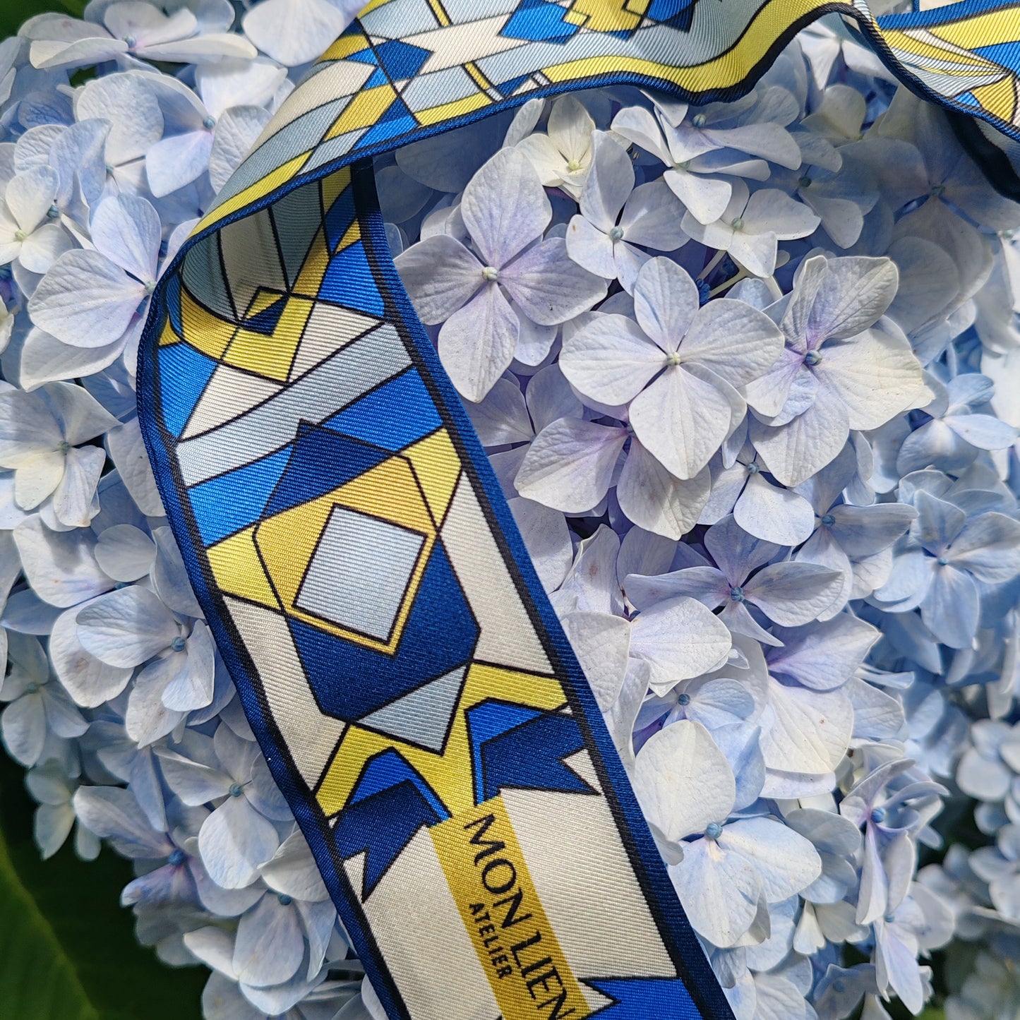 close up twilly in seta stampata con logo Mon Lien Atelier colore blu avorio giallo sopra fiore con petali azzurri